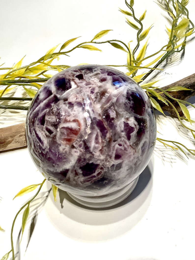 Pu&ograve; includere: Una grande sfera di ametista con un motivo a spirale di viola e bianco. La sfera &egrave; appoggiata su un piedistallo bianco.