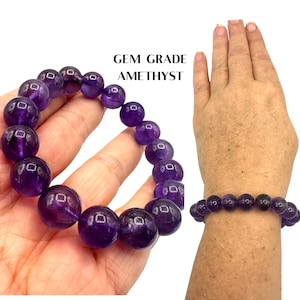 May include: A purple amethyst gemstone bracelet with round beads. The bracelet is shown on a person's wrist and hand.