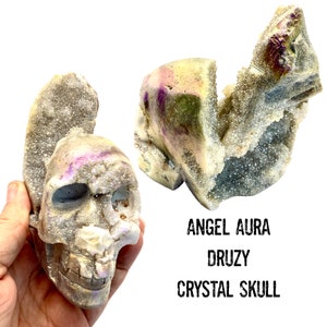 Puede incluir: Calavera de cristal Angel Aura Druzy con tonos iridiscentes de púrpura, dorado y verde. La calavera está tallada intrincadamente y presenta una superficie cristalina texturizada. La imagen también incluye el texto "ANGEL AURA DRUZY CRYSTAL SKULL."