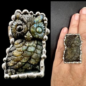 Peut inclure: Une bague imposante ornée d'une chouette sculptée en labradorite, sertie dans une monture en métal argenté à bord festonné. La sculpture présente des reflets irisés bleus et verts sur ses plumes texturées et ses yeux ronds, entourée de petites sphères argentées lisses sur tout le pourtour.