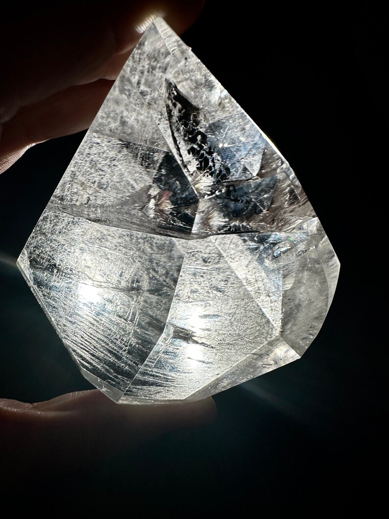 May include: A clear, faceted quartz crystal with internal striations and inclusions. The crystal is held against a black background, highlighting its geometric shape and transparency. The light catches the facets, creating a sparkling effect.