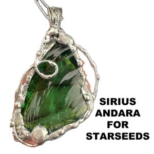May include: A handcrafted pendant featuring a large, irregular-shaped, green glass stone encased in a silver-colored metal frame. The pendant hangs from a delicate silver chain. The text "SIRIUS ANDARA FOR STARSEEDS" is displayed in black.