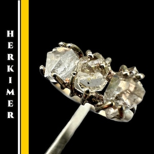 May include: A silver ring featuring three rough-cut, clear Herkimer diamond crystals. The crystals are set in a pronged design, with the word "HERKIMER" vertically on the left side of the image.