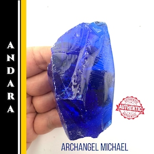 Puede incluir: Un cristal azul profundo, grande e irregular. El cristal se sostiene en una mano sobre un fondo blanco. El texto "ARCÁNGEL MICHAEL" está en la parte inferior, con sellos "AUTÉNTICO".