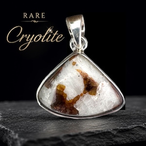 Peut inclure: Un pendentif triangulaire composé d'une pierre blanche et translucide avec des inclusions minérales brunes, serti dans une monture en métal argenté avec une bélière. Le texte RARE Cryolite est visible dans le coin supérieur gauche.