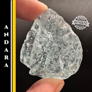 Puede incluir: Cristal Andara translúcido azul claro con una superficie rugosa y texturizada. El cristal se sostiene entre dos dedos. La imagen incluye la palabra "ANDARA" en texto blanco sobre un fondo negro y amarillo, y un sello blanco "AUTHENTIC".
