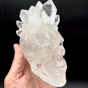 May include: A clear, translucent quartz crystal carved into the shape of a human skull. The top of the skull features a cluster of natural, pointed crystal formations extending upwards, with one prominent dark inclusion visible within the largest point.
