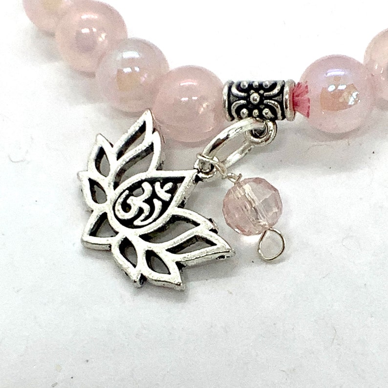 May include: A pink rose quartz beaded bracelet with a silver charm featuring a lotus flower and the Om symbol. The bracelet also includes a silver spacer bead and a pink crystal bead.