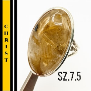 May include: A silver ring featuring a large, oval-shaped gemstone with golden, needle-like inclusions. The ring is size 7.5. The word "CHRIST" is printed vertically on the left side of the image.
