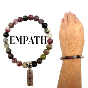 Puede incluir: Un brazalete hecho de cuentas de piedras preciosas de color rosa, verde y negro con un dije de plata que dice "BEAUTIFUL". El brazalete se muestra en la muñeca de una persona.