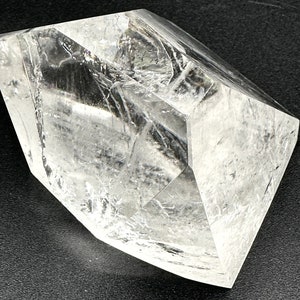 May include: A large, clear quartz crystal with multiple facets. The crystal is transparent, allowing light to pass through, and has a geometric shape. The surface appears to have internal fractures, adding texture. The crystal sits on a dark surface.