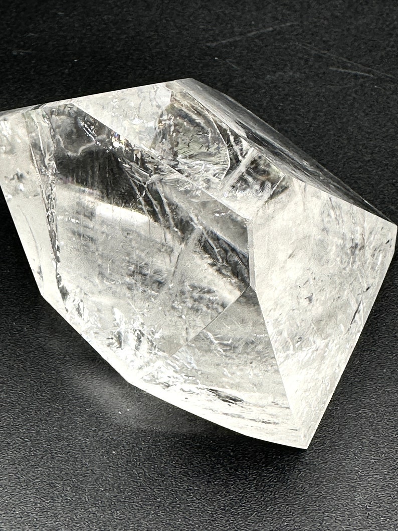 May include: A large, clear quartz crystal with multiple facets. The crystal is transparent, allowing light to pass through, and has a geometric shape. The surface appears to have internal fractures, adding texture. The crystal sits on a dark surface.