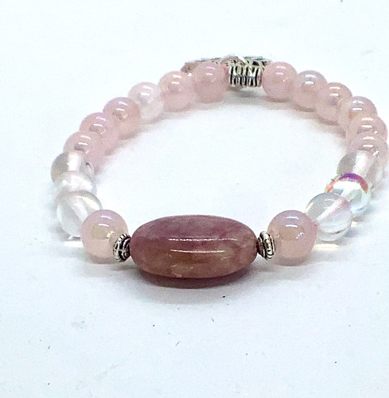 May include: A pink and white beaded bracelet with a larger oval-shaped pink stone in the center. The bracelet has a silver clasp.