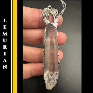 May include: A clear quartz crystal pendant with a silver-colored metal setting and chain. The pendant has a raw, natural appearance with visible internal lines. The setting features an openwork design. The word "LEMURIAN" is printed vertically on the left side.