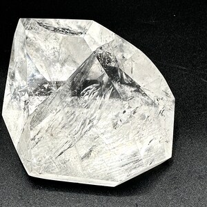 May include: A clear, faceted crystal with a geometric shape. The crystal has a polished surface with internal lines and inclusions, reflecting light. The crystal is set against a dark background, highlighting its clarity and form.