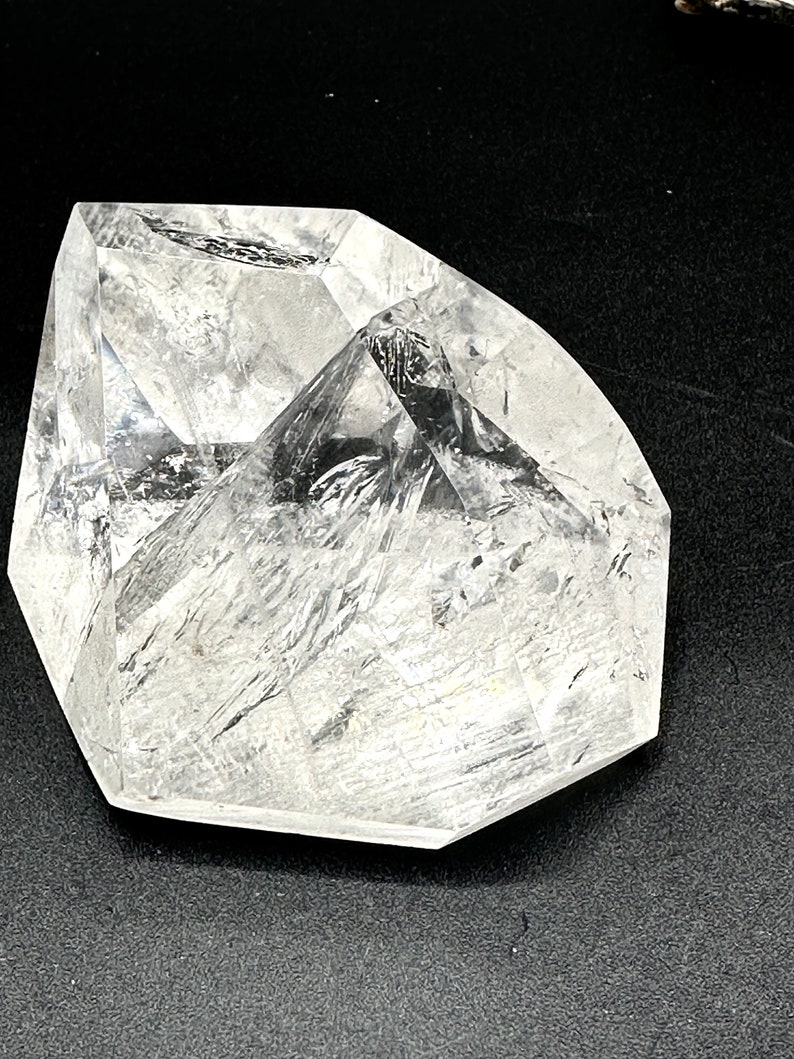 May include: A clear, faceted crystal with a geometric shape. The crystal has a polished surface with internal lines and inclusions, reflecting light. The crystal is set against a dark background, highlighting its clarity and form.