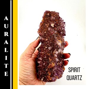 May include: Auralite Spirit Quartz crystal cluster. The crystal is a deep purple color with reddish-brown and white accents. The text "AURALITE" is vertically aligned on the left side of the image, and "SPIRIT QUARTZ" is on the right.