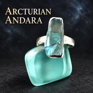 May include: A silver ring featuring a vertical, translucent light blue crystal set in a metal band. The ring rests on a smooth, rounded, opaque teal stone. The text ARCTURIAN ANDARA is displayed in the upper left corner against a dark background.