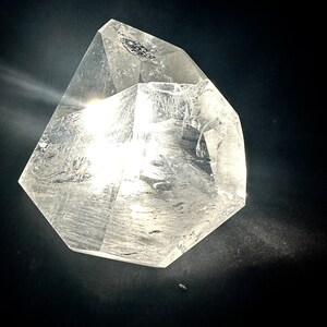 May include: A clear quartz crystal with a multifaceted, geometric shape. The crystal is transparent, allowing light to pass through, and is set against a dark background. The crystal's edges are well-defined, and it has a polished appearance.