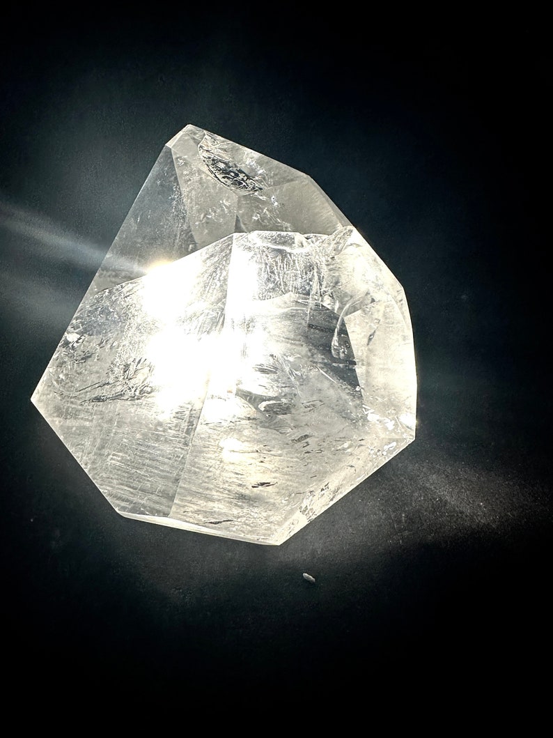 May include: A clear quartz crystal with a multifaceted, geometric shape. The crystal is transparent, allowing light to pass through, and is set against a dark background. The crystal's edges are well-defined, and it has a polished appearance.