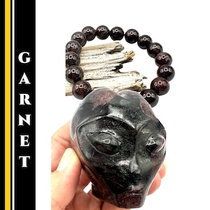 May include: A dark red garnet alien head sculpture and a garnet bead bracelet. The sculpture is held in a hand and has carved facial features. The bracelet is strung on a cord and has round beads. The word "GARNET" is written vertically in white letters.