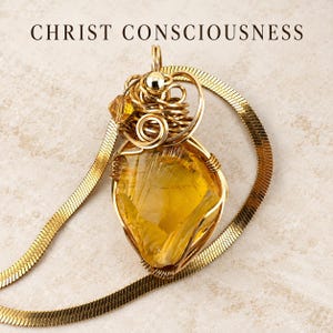 May include: A gold-toned pendant featuring a large, translucent yellow gemstone encased in intricate gold wire wrapping, suspended from a flat gold herringbone chain. The text CHRIST CONSCIOUSNESS is printed in black above the jewellery piece.