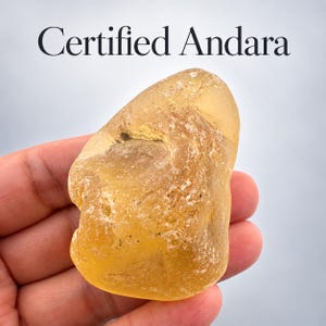 May include: A translucent, irregular yellow Andara crystal with a smooth, polished surface and visible internal textures. The text "Certified Andara" is printed in black at the top of the image, identifying the yellow, glass-like mineral specimen.