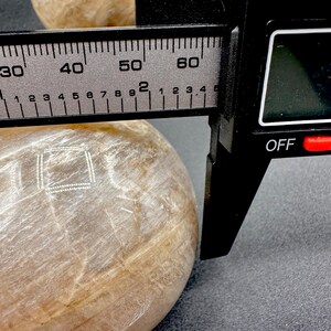 Pode incluir: Uma esfera de pedra redonda, lisa, polida, branca e transl&uacute;cida com um desenho quadrado branco na superf&iacute;cie. A esfera est&aacute; sendo medida com um paqu&iacute;metro preto. O paqu&iacute;metro indica 50 mm.