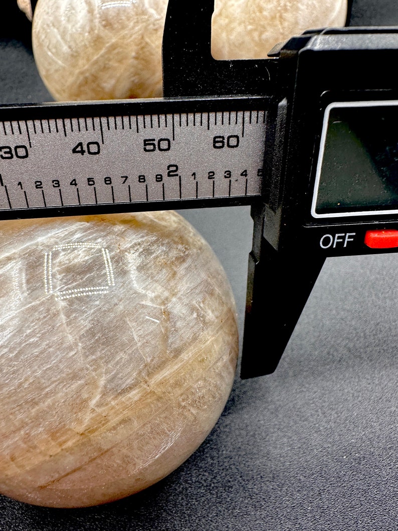 Pode incluir: Uma esfera de pedra redonda, lisa, polida, branca e transl&uacute;cida com um desenho quadrado branco na superf&iacute;cie. A esfera est&aacute; sendo medida com um paqu&iacute;metro preto. O paqu&iacute;metro indica 50 mm.