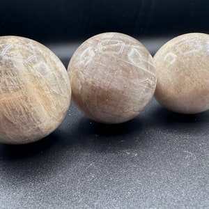 Pode incluir: Tr&ecirc;s esferas lisas, brancas e marrons de cristal de selenita. As esferas est&atilde;o alinhadas em uma fila sobre uma superf&iacute;cie preta.