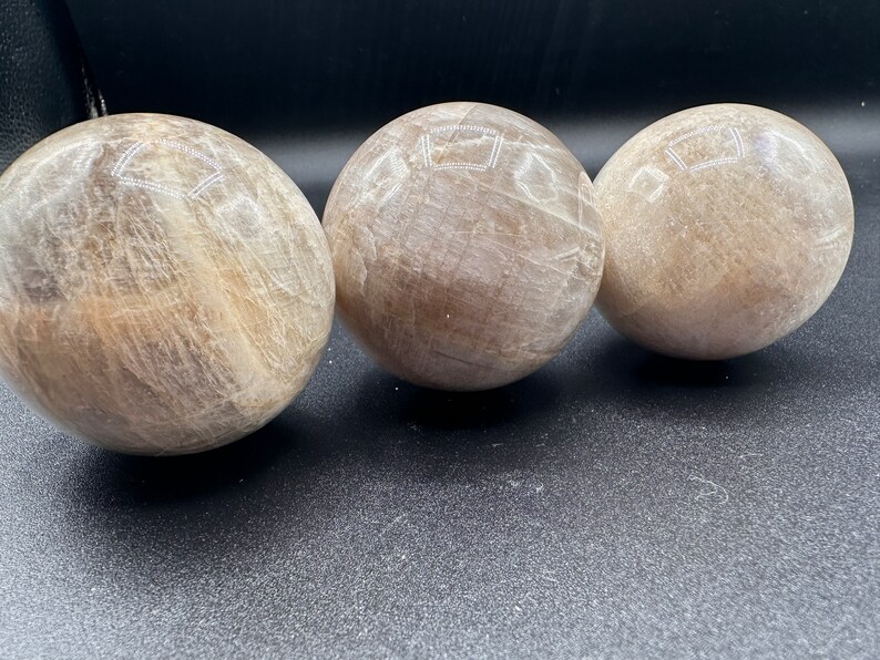 Pode incluir: Tr&ecirc;s esferas lisas, brancas e marrons de cristal de selenita. As esferas est&atilde;o alinhadas em uma fila sobre uma superf&iacute;cie preta.