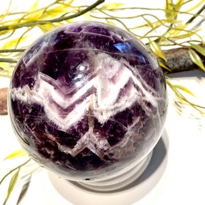 Pu&ograve; includere: Una sfera di ametista viola con inclusioni di cristallo bianco. La sfera &egrave; lucidata e ha una superficie liscia e lucente.