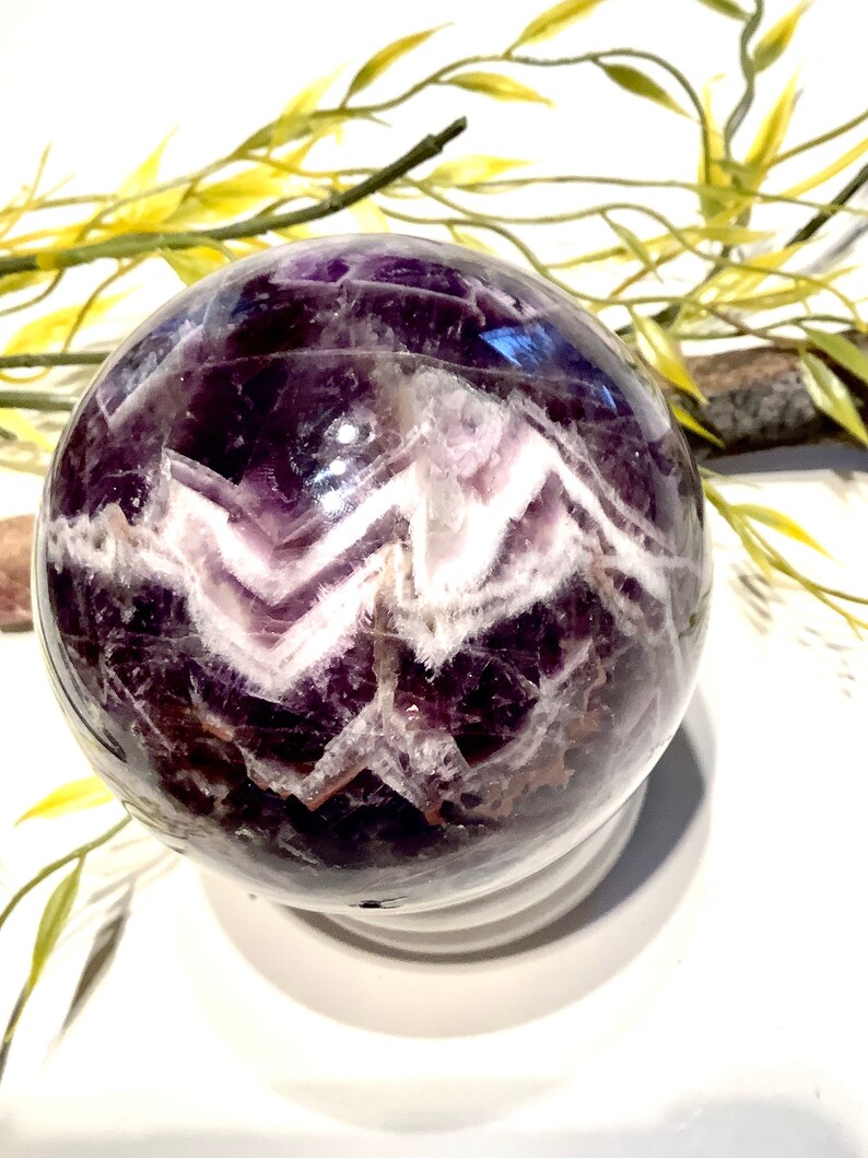 Pu&ograve; includere: Una sfera di ametista viola con inclusioni di cristallo bianco. La sfera &egrave; lucidata e ha una superficie liscia e lucente.