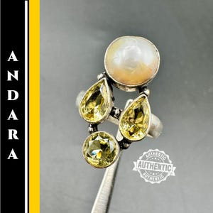 May include: A silver ring featuring a large, round, off-white pearl and three yellow-green gemstones. The ring has a unique design with the gemstones arranged in a cluster. The word "ANDARA" is printed vertically on the left side of the image.