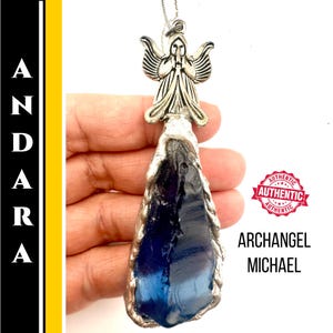 Puede incluir: Un collar con colgante plateado con una figura de ángel sobre una piedra azul oscuro de forma irregular. El colgante cuelga de una fina cadena. Las palabras "ARCHANGEL MICHAEL" están impresas en negro.
