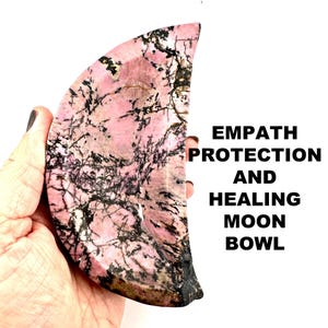 May include: A pink and black moon-shaped bowl made of polished gemstone. The bowl is carved from a single piece of stone and has a natural, organic shape. The text "Empath Protection and Healing Moon Bowl" is printed in black on a white background.