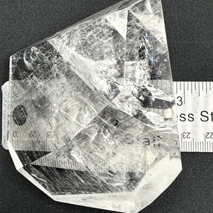 May include: A clear quartz crystal with a faceted, geometric shape. The crystal is transparent, allowing light to pass through, and has internal fractures. A ruler is placed next to the crystal for scale, showing it is approximately 3 inches long.