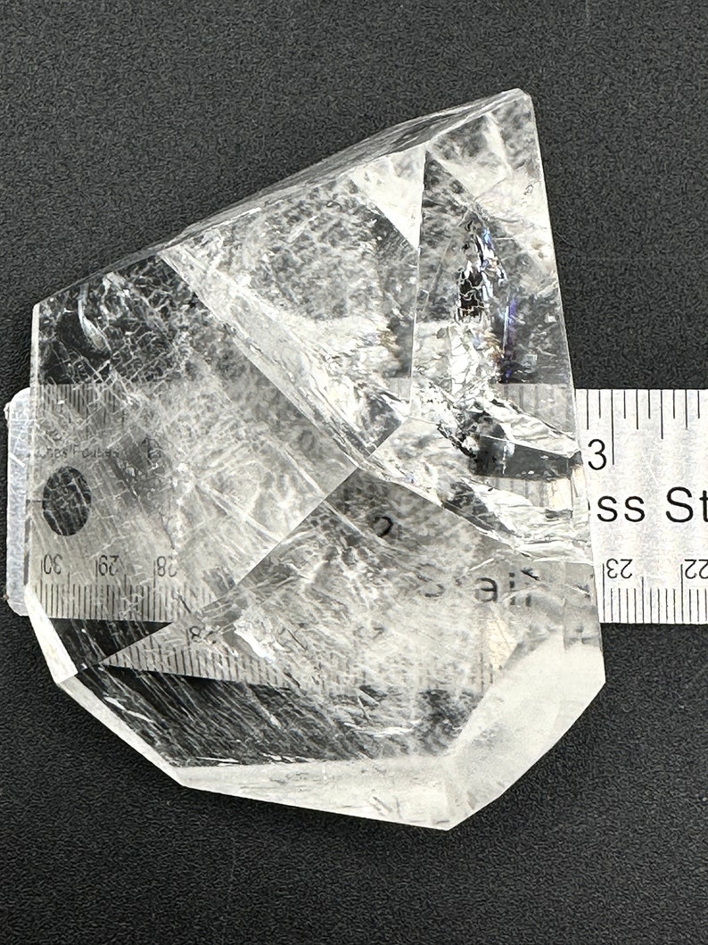 May include: A clear quartz crystal with a faceted, geometric shape. The crystal is transparent, allowing light to pass through, and has internal fractures. A ruler is placed next to the crystal for scale, showing it is approximately 3 inches long.
