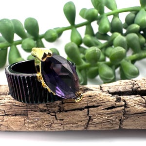Puede incluir: Un anillo con una banda ancha de color negro con estrías verticales y una gran piedra preciosa de color púrpura en talla marquesa, engastada en un marco de metal dorado. La piedra púrpura facetada destaca sobre la banda oscura.