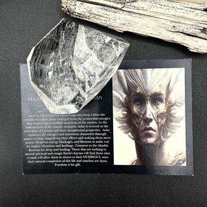 May include: A clear quartz crystal with a faceted shape, resting on a black surface next to a card with an illustration of a person with a detailed face and text. A piece of driftwood is in the background.
