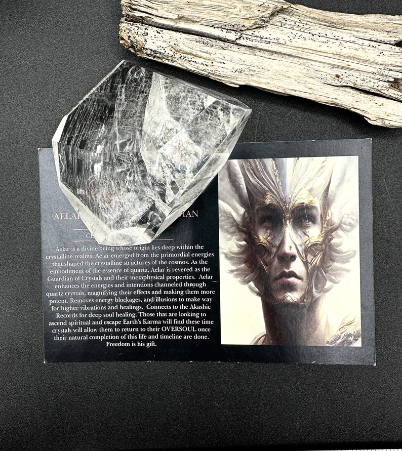 May include: A clear quartz crystal with a faceted shape, resting on a black surface next to a card with an illustration of a person with a detailed face and text. A piece of driftwood is in the background.