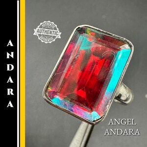 Puede incluir: Un anillo de plata con una gran gema roja rectangular con bordes iridiscentes azules y verdes. El anillo está sobre un fondo oscuro. El texto "ANGEL ANDARA" es visible.