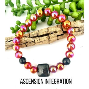 Può includere: Un braccialetto di perline con perline rotonde iridescenti nei toni del rosso, dell'arancione e del viola, intervallate da perline nere e una pietra quadrata grigio scuro. Il testo "ASCENSION INTEGRATION" è visualizzato sotto.
