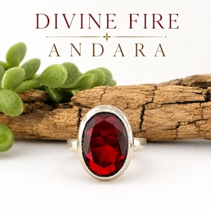 May include: A silver ring featuring a large, oval-cut, deep red gemstone. The text DIVINE FIRE ANDARA is printed in dark red and gold above the ring. The ring is set against a background of a piece of textured wood and green succulent leaves.