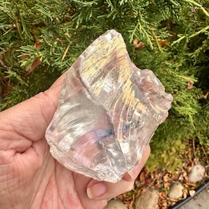 May include: A large, irregular chunk of clear, translucent glass with a smooth, polished surface. The glass features subtle iridescent highlights that catch the light, creating a reflective, multi-faceted appearance with curved, shell-like fracture patterns throughout the material.