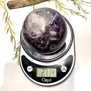 Pu&ograve; includere: Una grande sfera di cristallo di ametista che pesa 2,714 libbre su una bilancia digitale. La sfera &egrave; di colore viola scuro con inclusioni bianche e trasparenti.