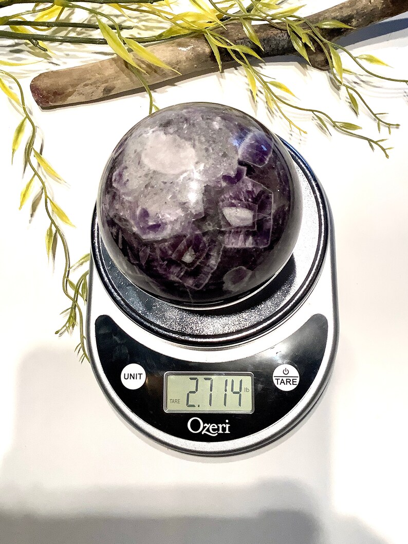 Pu&ograve; includere: Una grande sfera di cristallo di ametista che pesa 2,714 libbre su una bilancia digitale. La sfera &egrave; di colore viola scuro con inclusioni bianche e trasparenti.