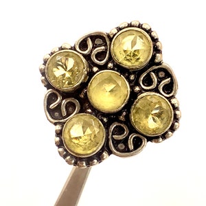May include: A silver-toned ring featuring a floral design with six round, faceted, pale yellow gemstones. The ring has an ornate, openwork design with a beaded border and is held by tweezers against a white background.