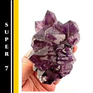 May include: A cluster of deep purple Super 7 crystals, held in a hand, with a white background. The crystals have sharp, geometric points and a rough, textured surface. The text "SUPER 7" is visible on the left side of the image.