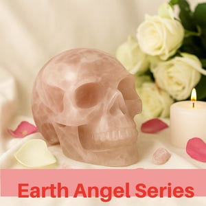 May include: A carved rose quartz skull, a symbol of transformation, sits on a white surface. The skull is a pale pink color, with visible details of the bone structure. The text "Earth Angel Series" is at the bottom of the image.